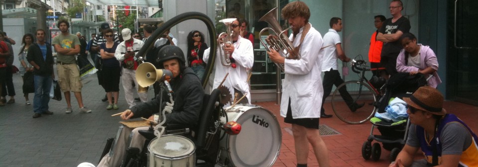 Man wearing crash helmet in electric wheelchair with drums attached to it, followed by two men in lab coats and shorts playing tubas in a plaza outside a bus station